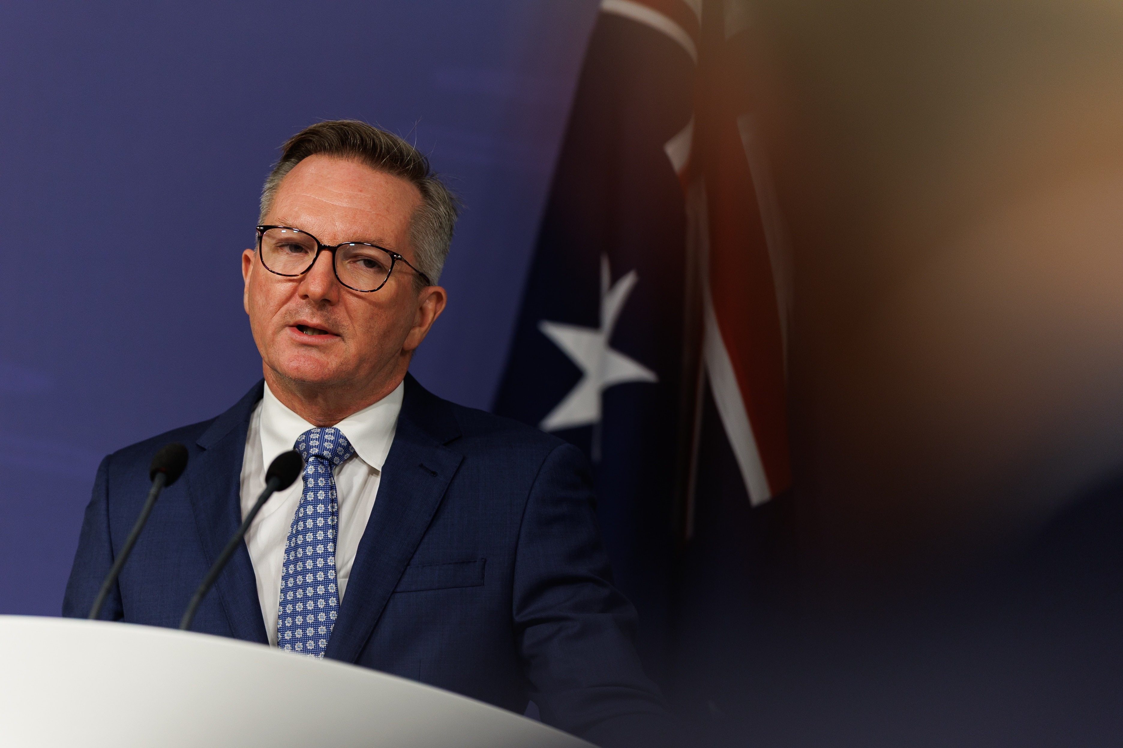 Chris Bowen looks slightly fed up as he stands in front of an Australian flag at a press conference.