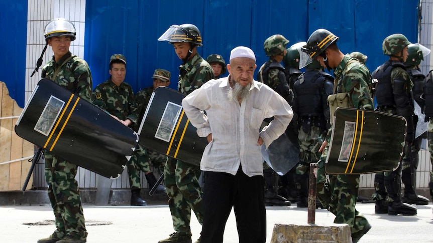 A man holds his hands on his hips as soldiers walk behind him.