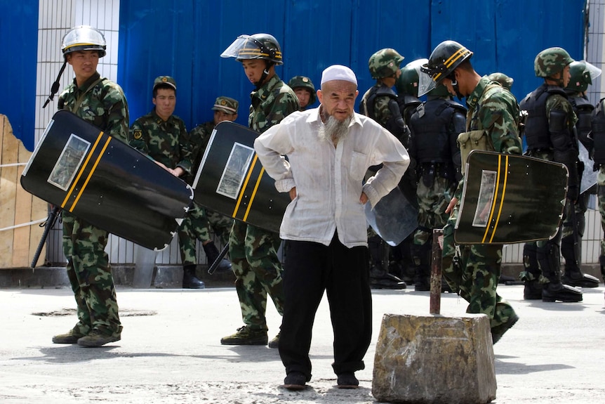 A man holds his hands on his hips as soldiers walk behind him.