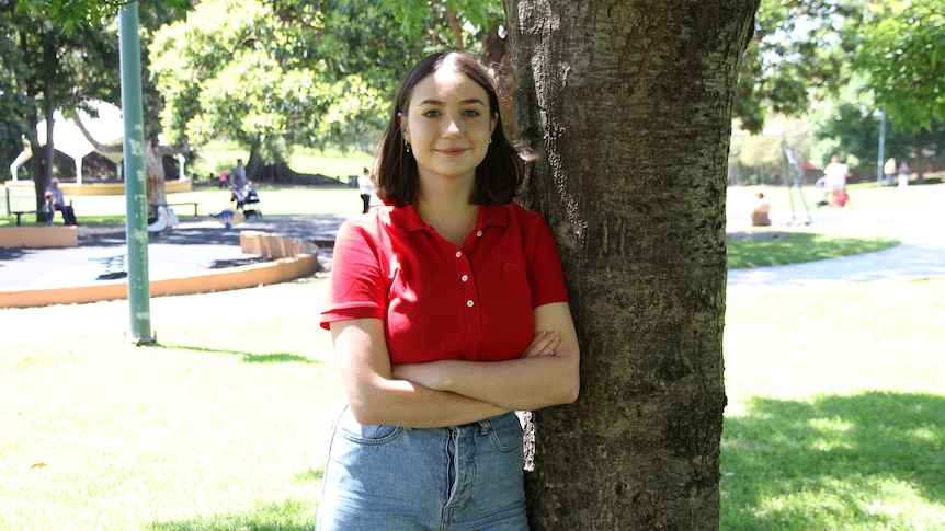 A woman leans against a tree in a red shirt.
