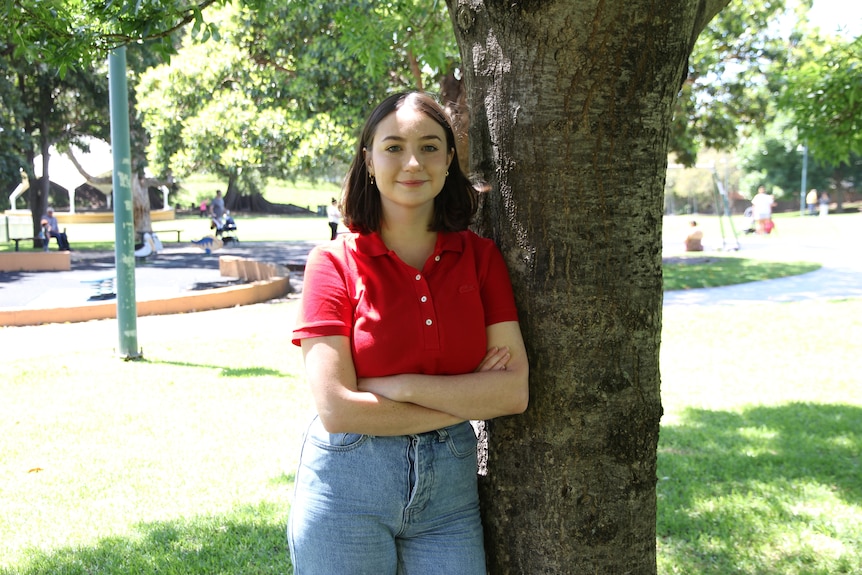 A woman leans against a tree in a red shirt.