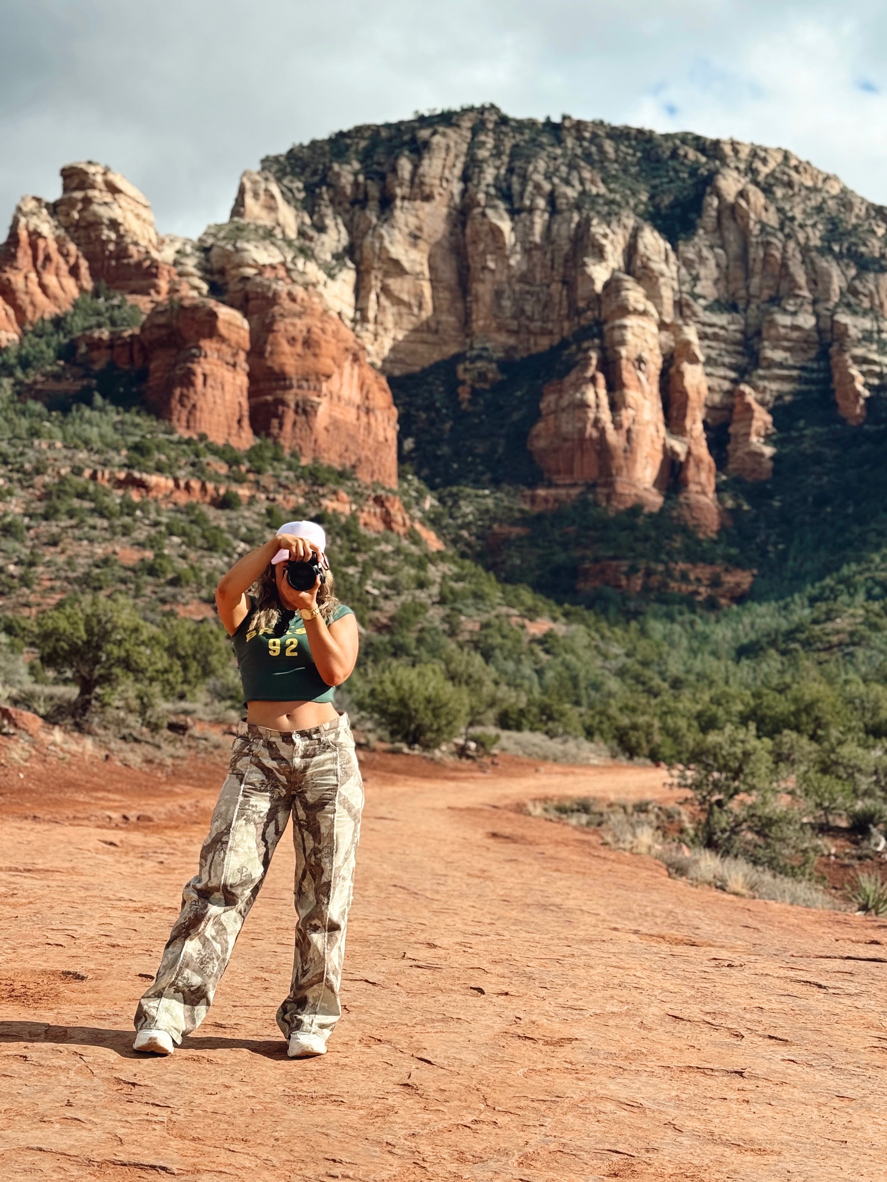 Bek stands in front rock formations takes a photo with camera, cropped top, cap, face hidden behind camera.
