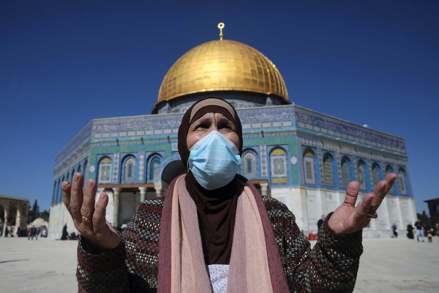 A woman in a veil and face mask with her arms outstretched while standing before a golden domed mosque