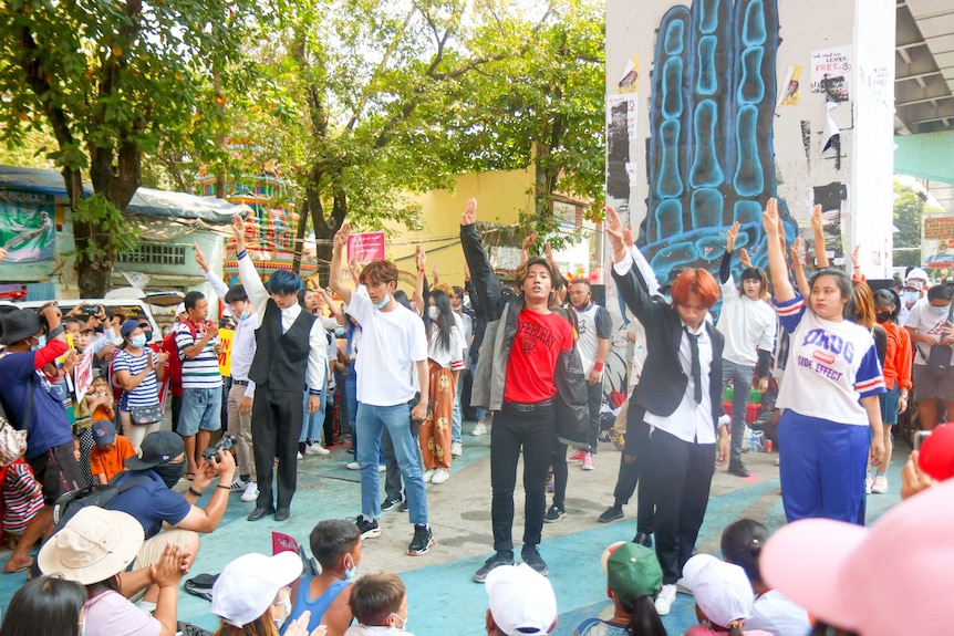 A group of young people holding up three fingers as they stand surrounded by others.