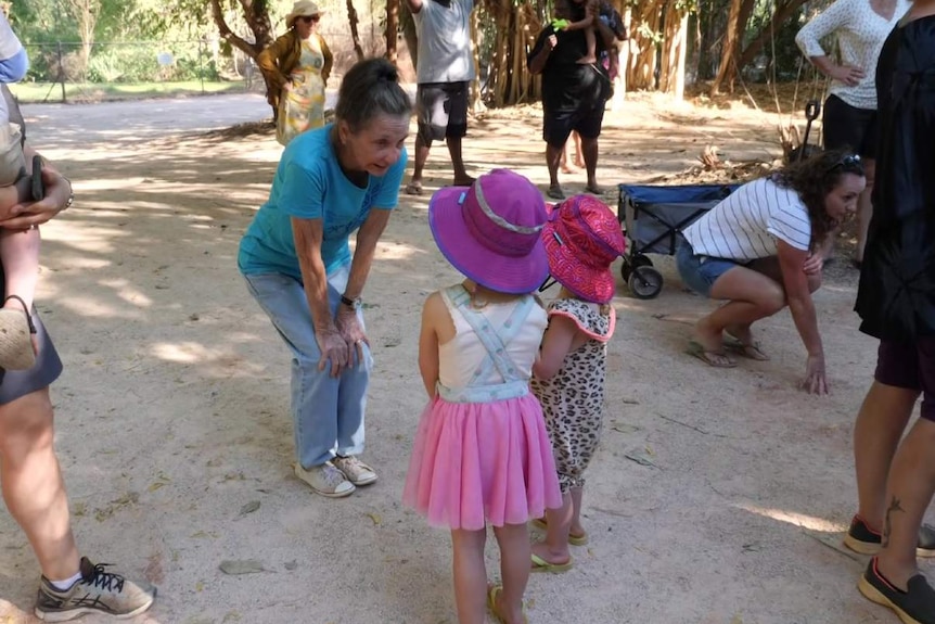 An older woman in a blue t-shirt leans over with her hands on her knees talking to two small children in a crowd.