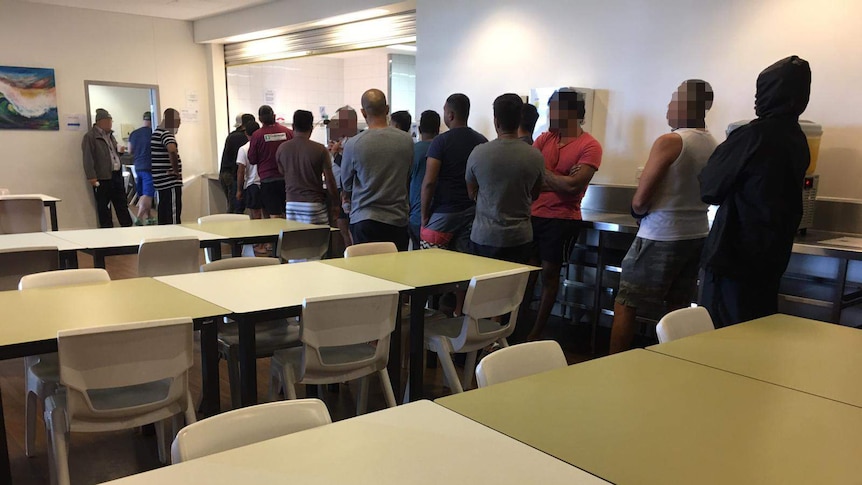 Men with faces pixilated stand in queue toward food service area with security guard standing nearby