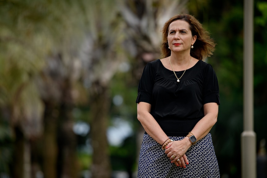 A woman wearing a black top and blue skirt stands in a park. Her hands are clasped as she looks to the distance.