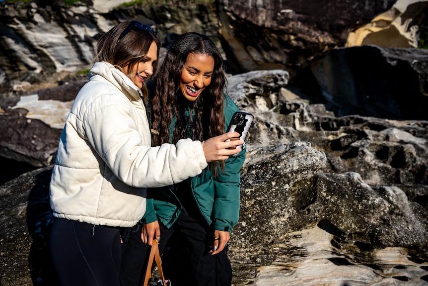 Woman wearing white jacket shows phone to woman in green jacket. 