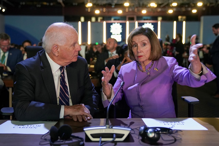 Nancy Pelosi sits at a desk with Ben Cardin, mid gesture. 