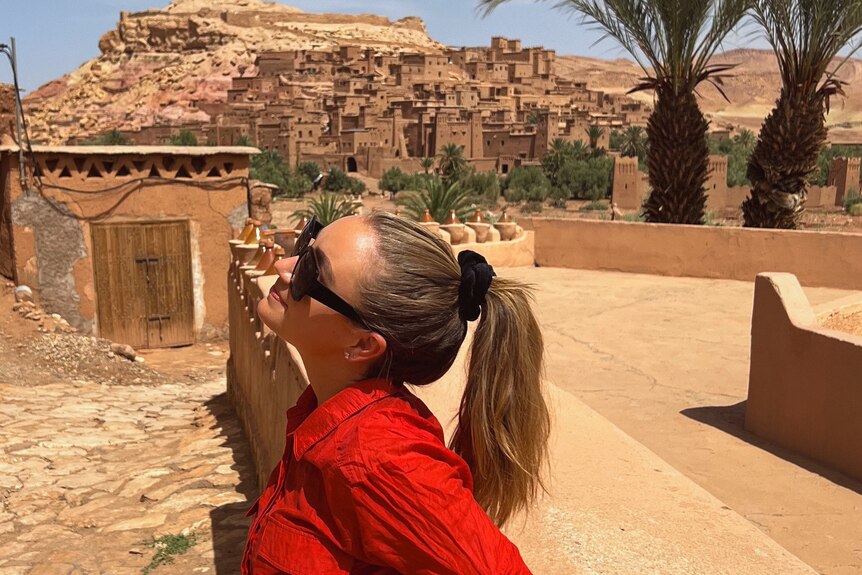 A lady standing sideways to the camera in a red shirt in Morocco with buildings on a hill behind her