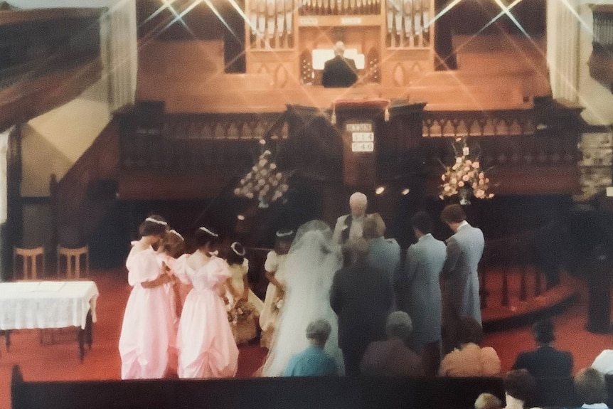 A coloured photo shows a bride in white and groomsmen in blue, bridesmaid in pink frothy dresses at the alter of an old church. 