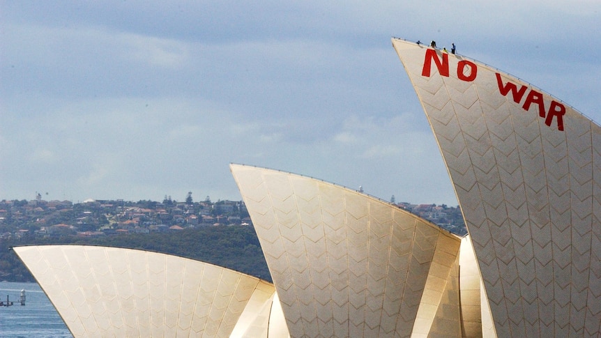 Three sails of the Sydney Opera House jut out above Sydney Harbour. Red letters spelling 'NO WAR' are painted on the main sail.
