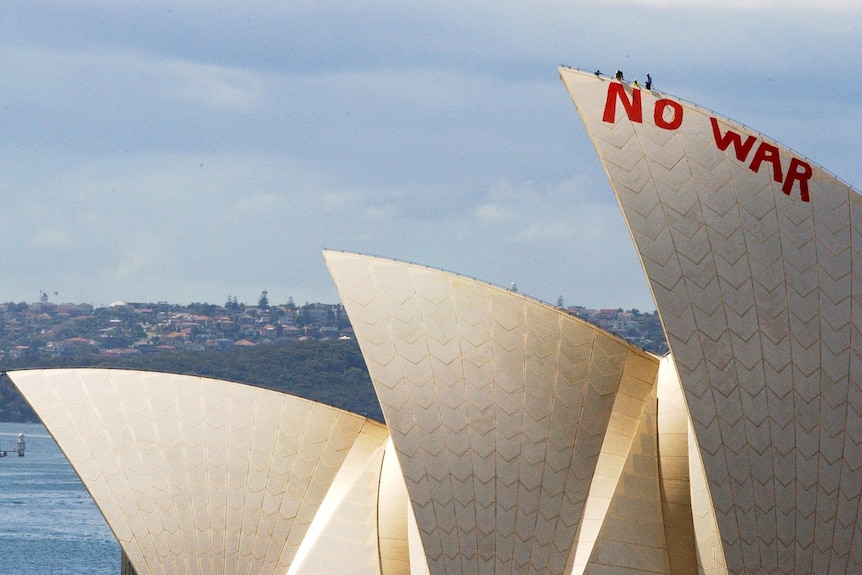Three sails of the Sydney Opera House jut out above Sydney Harbour. Red letters spelling 'NO WAR' are painted on the main sail.
