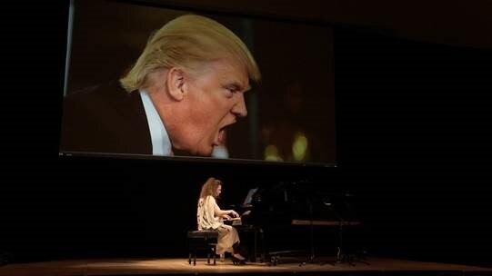 A pianist on a stage under a projector showing a large image of Trump speaking
