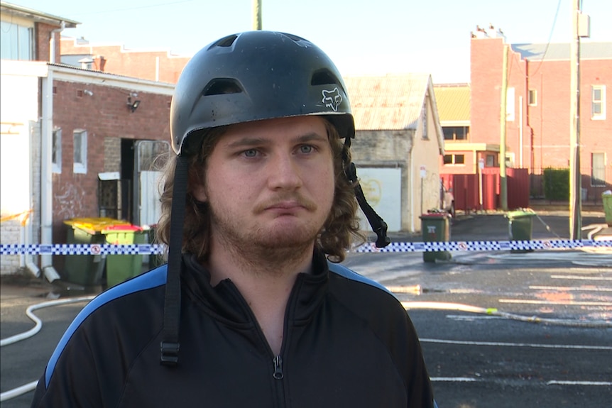 A young man with curly hair and a bike helmet on his head stands in front of police tape.