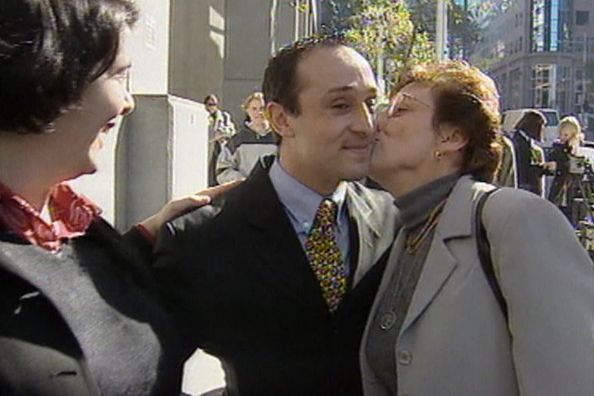 A man smiles as an older woman kisses him on the cheek. They are standing on the footpath, surrounded by other people.