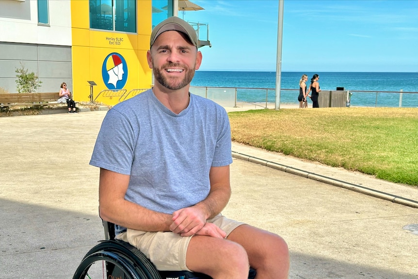 A man sits in a wheelchair smiling with the beach in the background