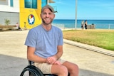 A man sits in a wheelchair smiling with the beach in the background