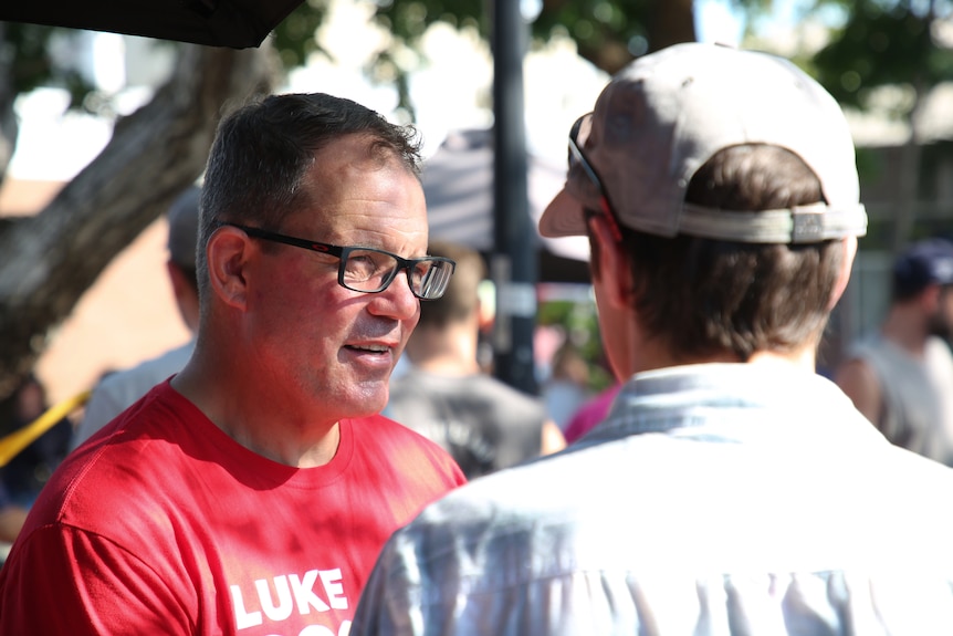 A man in a red shirt which reads 'Luke Gosling' looks serious as he speaks to another man.