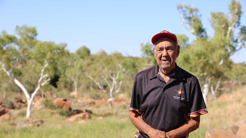 Senior Indigenous man wearing cap and polo shirt stands in outback landscape