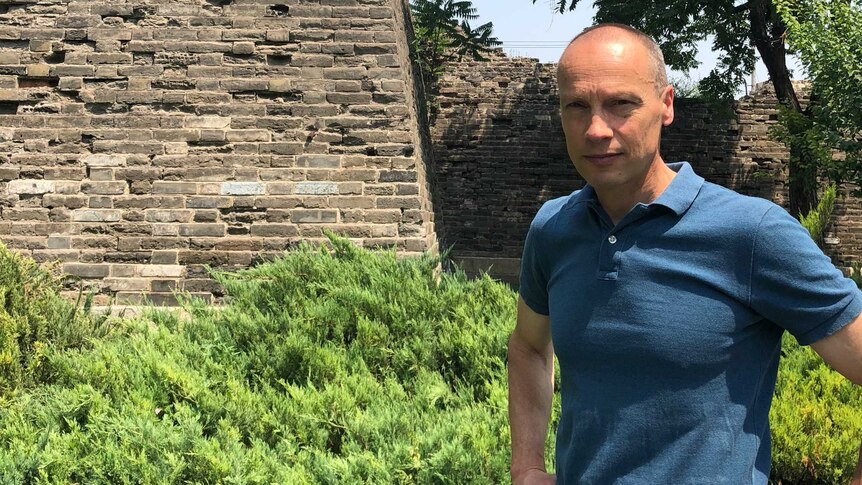 Man stands with hands on hips in front of a bluestone wall.