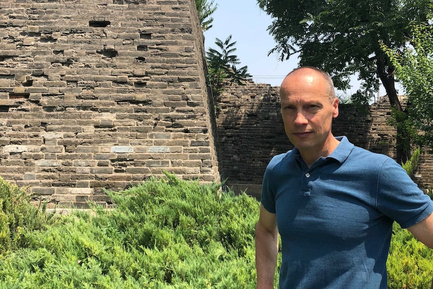 Man stands with hands on hips in front of a bluestone wall.