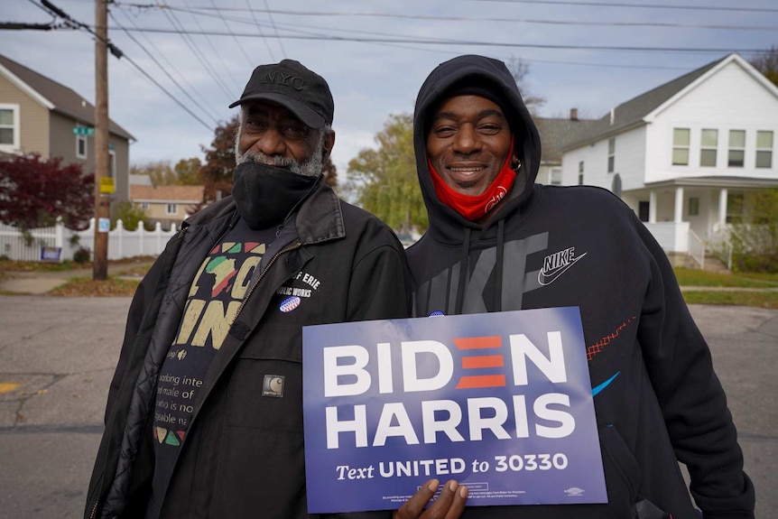 two men in black jumpers hold up a BIDEN HARRIS campaign sign while standing in a street outside