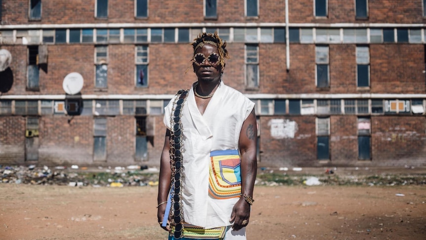 Blick wears a white shirt with a colourful pocket and sunglasses. He stands in front of a red brick apartment complex.