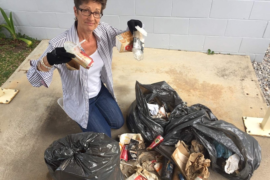 A woman sits next to four full garbage bags of rubbish and holds up some of their contents.