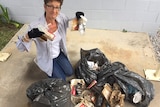 A woman sits next to four full garbage bags of rubbish and holds up some of their contents.