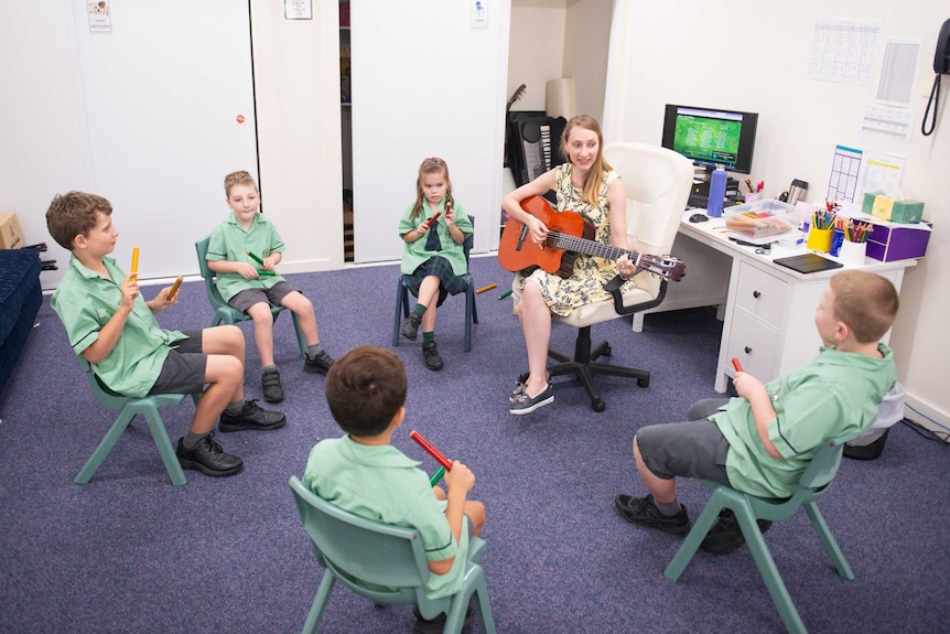 Young students take part in a music lesson with female teacher playing guitar.