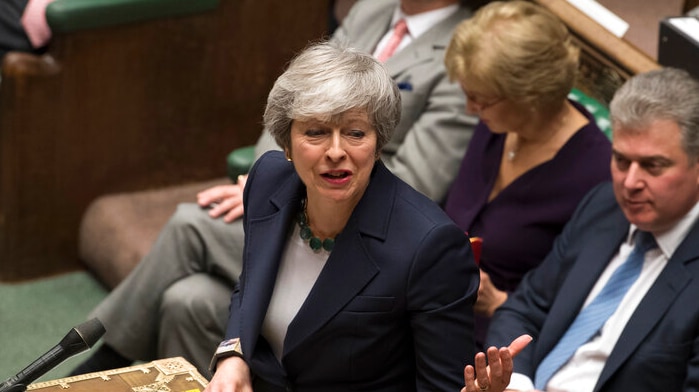 Theresa may standing at a parliamentary lectern, wearing a dark jacket. She is mid-speech.