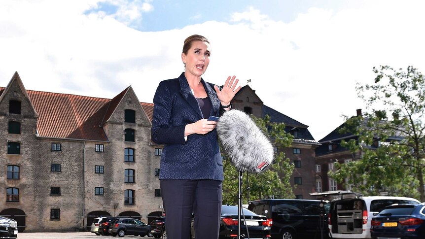 Denmark's Prime Minister Mette Frederiksen stands on a sunny street wearing a navy suit speaking into a microphone and gesturing