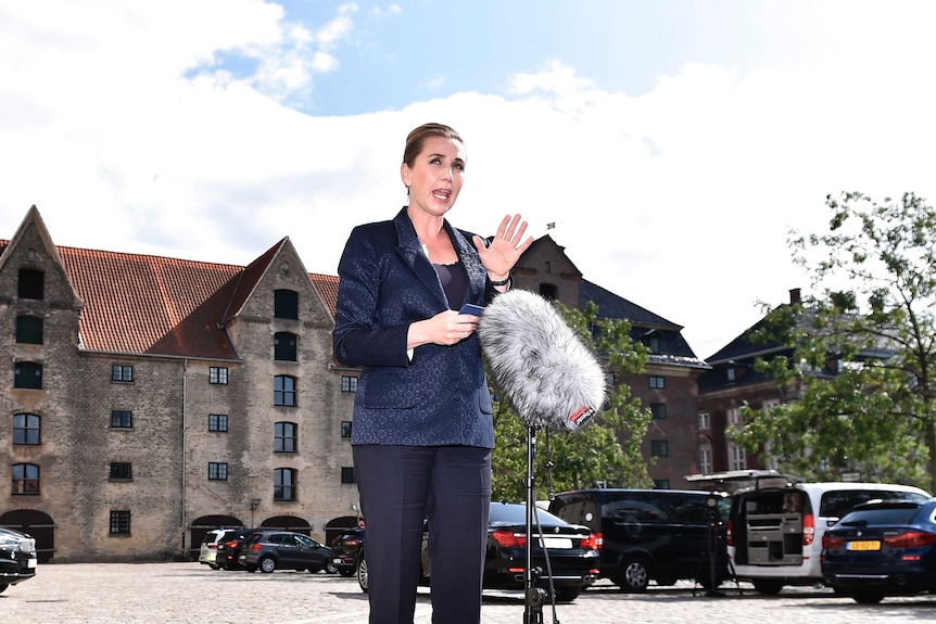 Denmark's Prime Minister Mette Frederiksen stands on a sunny street wearing a navy suit speaking into a microphone and gesturing