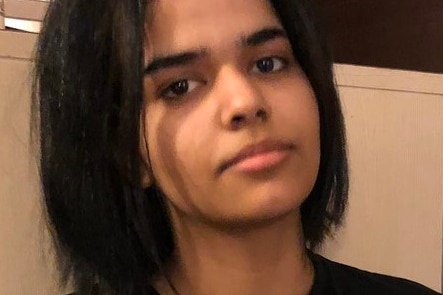 A young woman wearing a black t shirt and ripped jeans sits near a bed in a small hotel room.