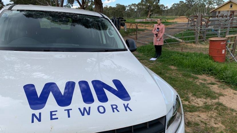 A white car with the words WIN Network on the bonnet. A young female journalist speaks into a microphone in the background