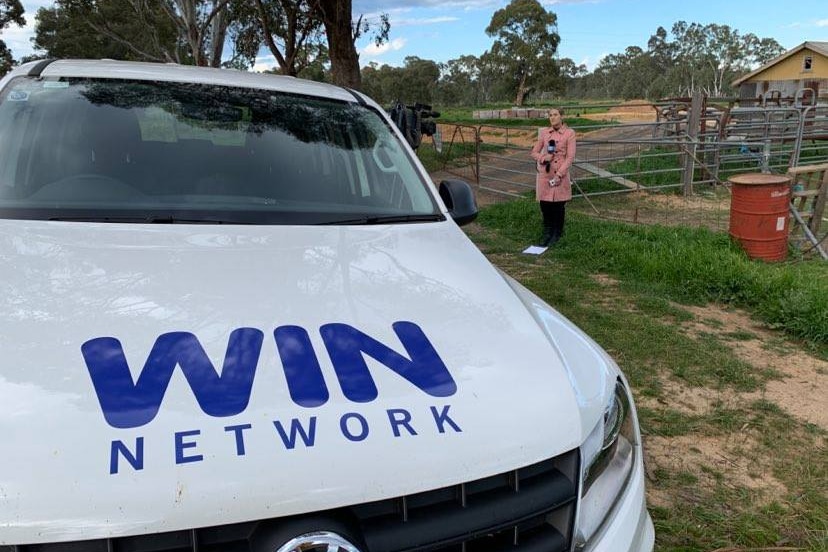 A white car with the words WIN Network on the bonnet. A young female journalist speaks into a microphone in the background