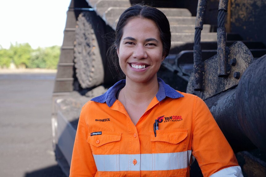 a young woman wearing hi-vis workwear stands in front of a mining truck, smiling at the camera