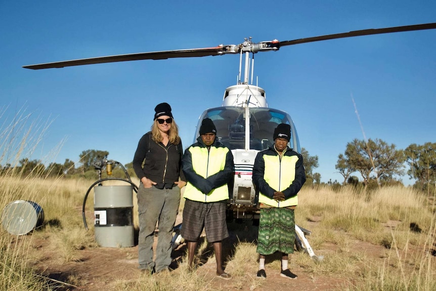 Three woman, two of them Aboriginal, stand in front of a helicopter on country