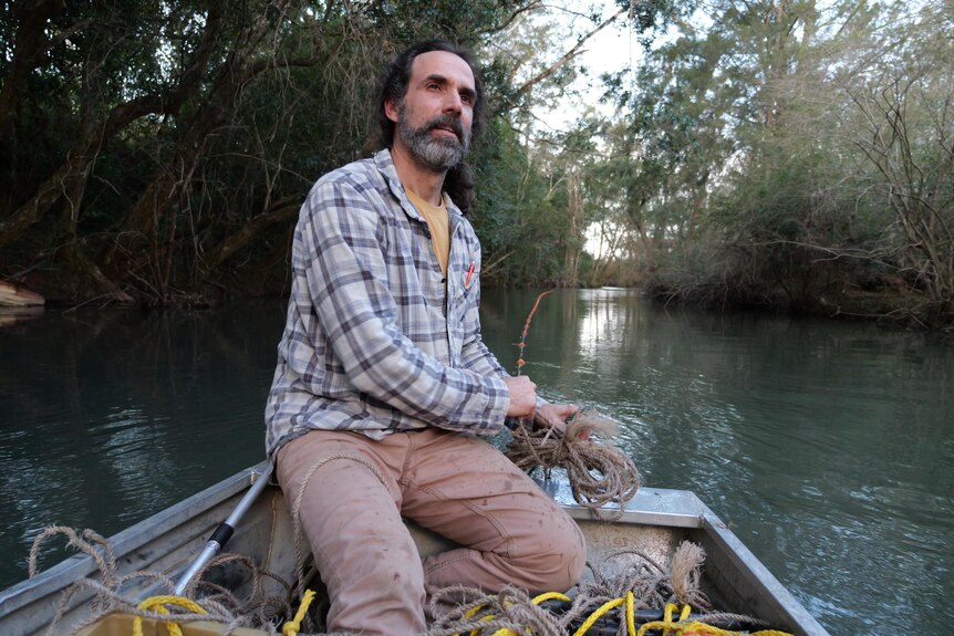 A man in a checked shirt and beard sits on a tin boat in a river.