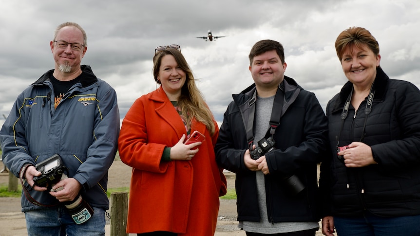 four people stand in a row smiling, they are holding cameras and a plane flies above them.
