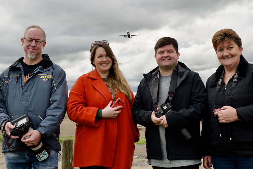 four people stand in a row smiling, they are holding cameras and a plane flies above them.
