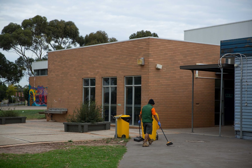 A groundskeeper wearing a fluoro shirt and vest picks up rubbish in the schoolyard