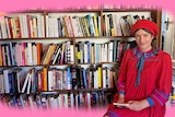 A woman in a red beret and dress sits in front of a large and very well stocked bookshelf