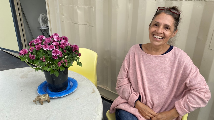 A woman sits grinning at the camera next to a table with potted flowers on it. A shipping container wall in background.