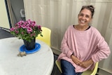 A woman sits grinning at the camera next to a table with potted flowers on it. A shipping container wall in background.