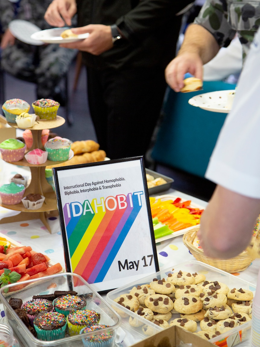 A range of baked goods on a table with people around it and a sign in the middle in rainbow saying IDAHOBIT