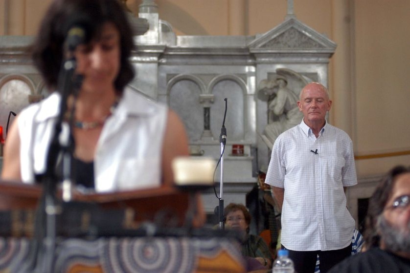 A woman delivers a homily in a church while a priest in his 70s stands behind her.