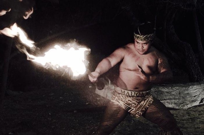 A shirtless man, half squatting, with fire in the nighttime, wearing a headband