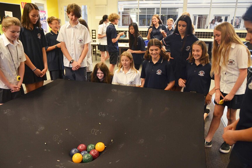 A wide shot of high school students in a classroom standing around a large black funnel with coloured balls in the middle.